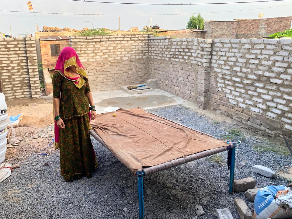A woman standing beside a charpoy in an unfinished brick courtyard.